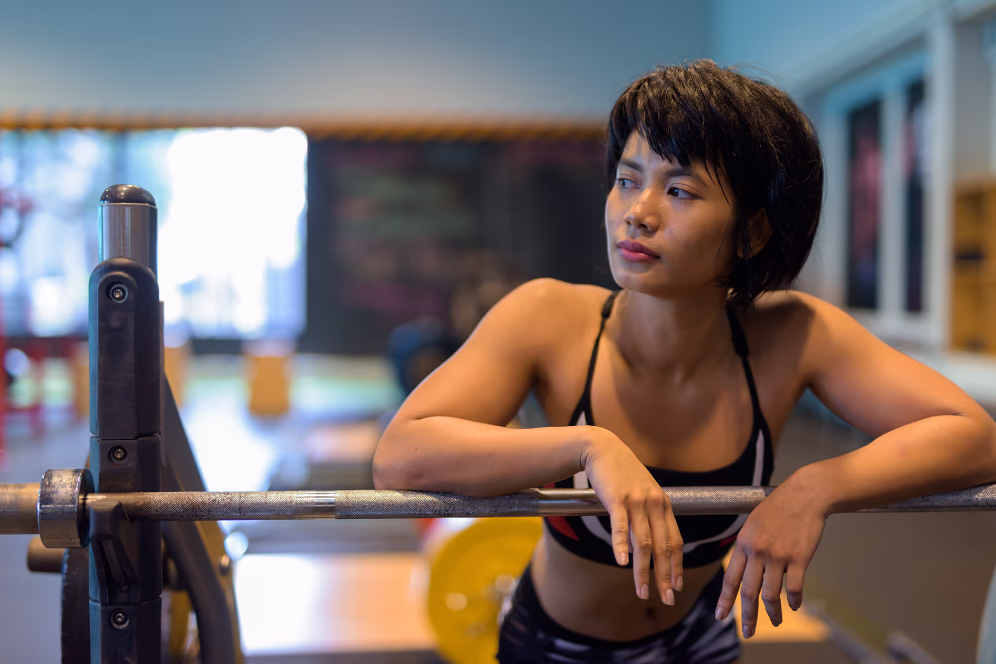 Young woman in sportswear leaning on a barbell in a gym, looking to the side.