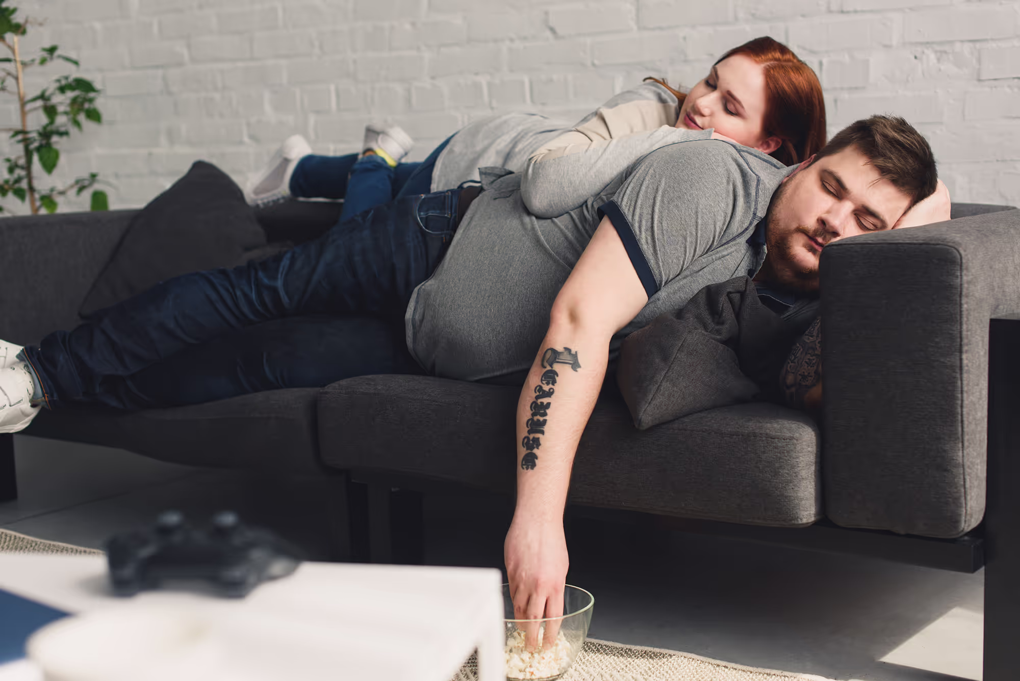Overweight couple sleeping on a gray couch, man resting his hand in a bowl of popcorn.