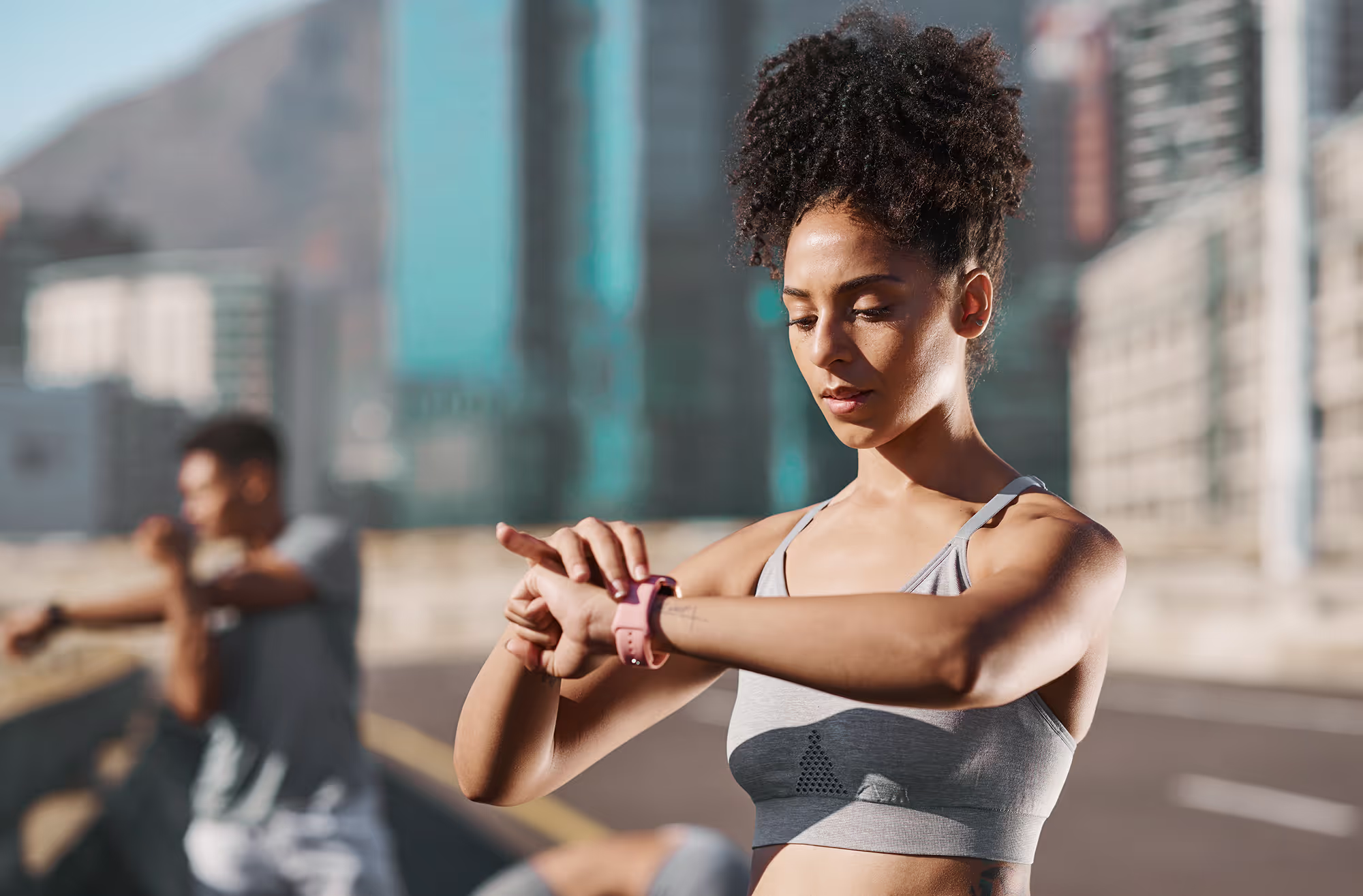 Athletic woman in a gray sports bra checking her smartwatch outdoors with a blurred person stretching in the background.