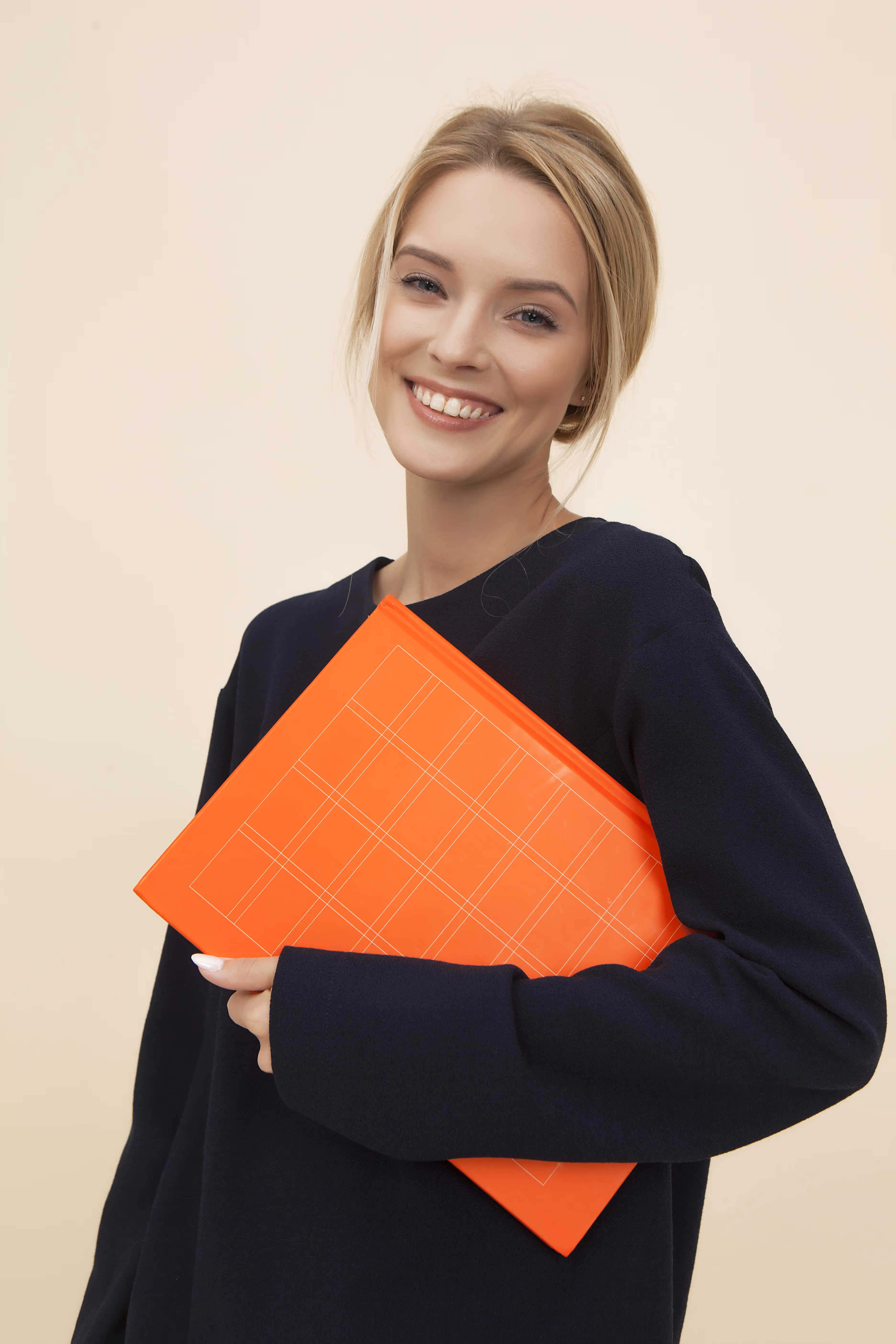 Smiling blonde woman in a black sweater holding an orange notebook against a beige background.