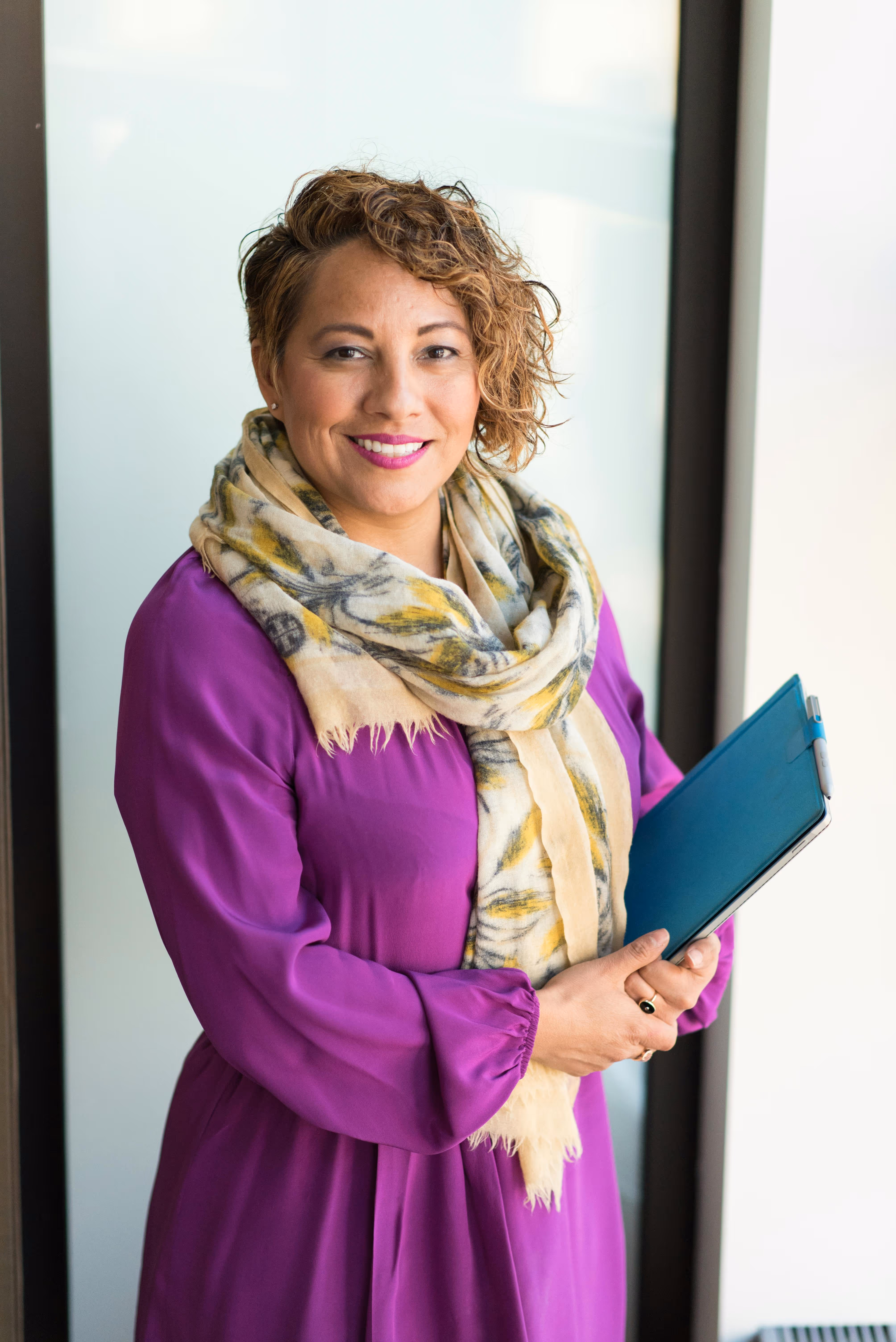 Smiling woman with curly hair wearing a purple dress and patterned scarf, holding a blue tablet.
