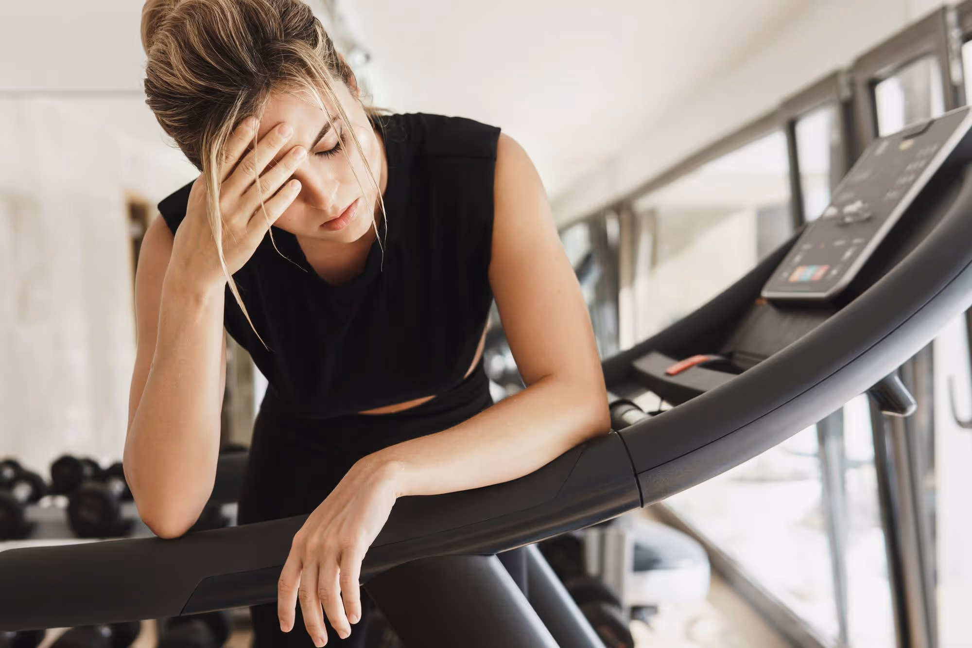 Tired woman in black workout clothes leaning on a treadmill, holding her forehead in a gym.