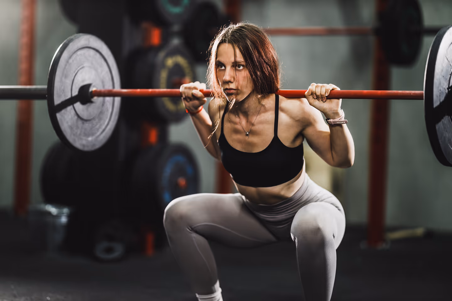 Woman in black sports bra and gray leggings performing a weighted squat with a barbell in a gym.