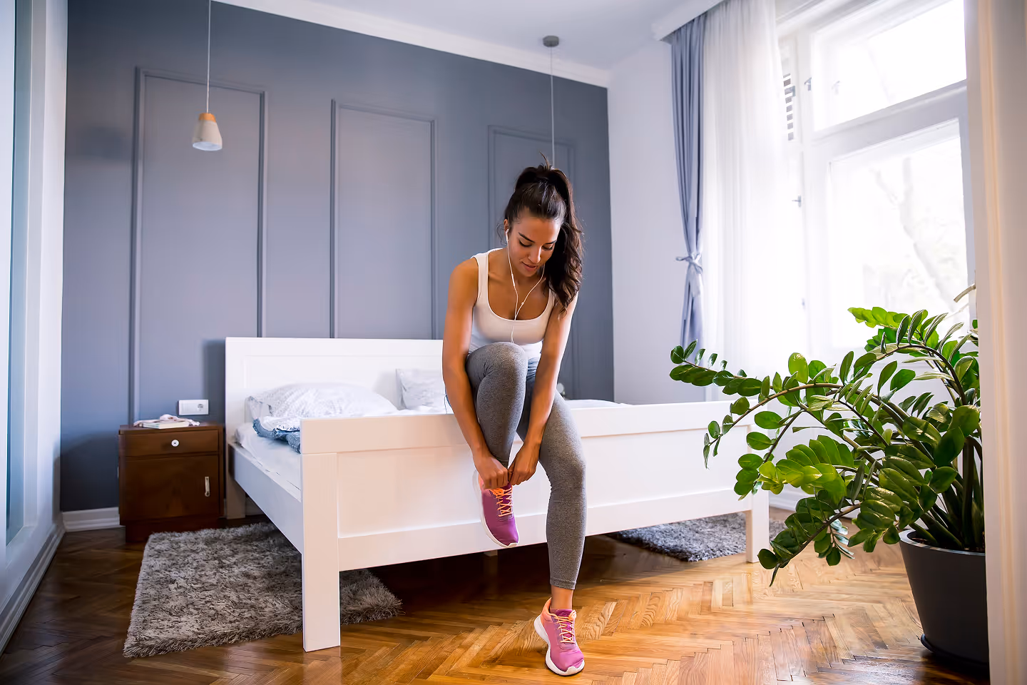 Young woman sitting on a white bed tying pink running shoes in a bright bedroom with wooden floor and a green plant.