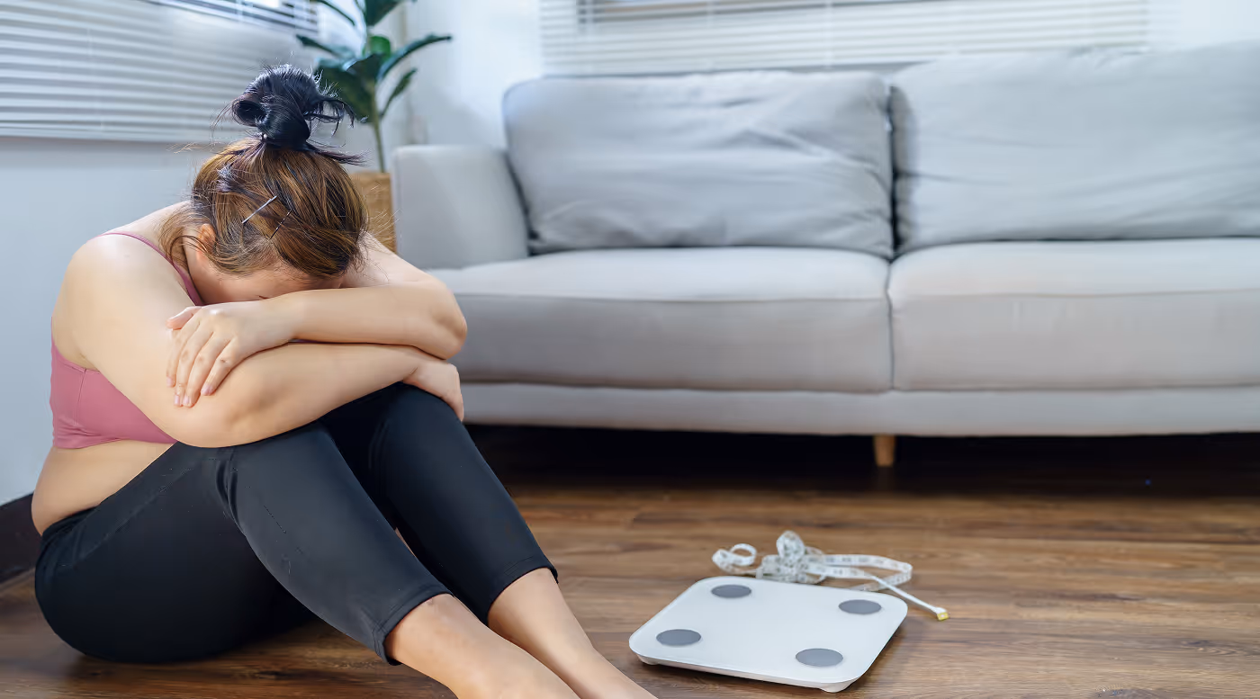 Woman sitting on the floor with head resting on folded arms, next to a scale and measuring tape on a wooden floor.